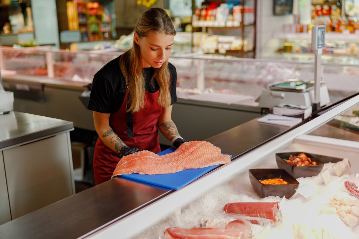 A fishmonger in a red apron and black gloves prepares a salmon fillet behind a display case of seafood.