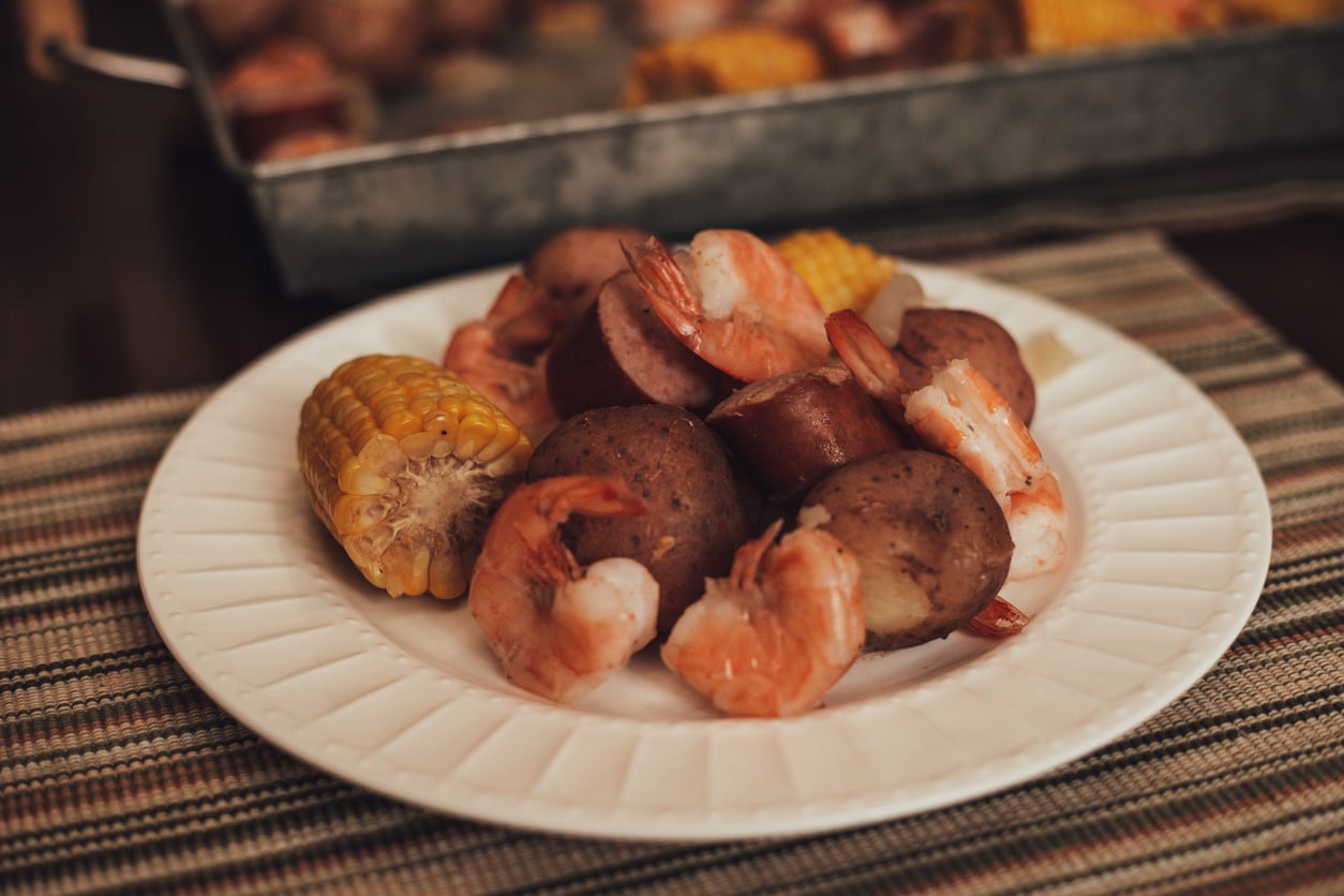A plate containing shrimp, sausage, and corn on the cob, with a larger tray of the same meal in the background.