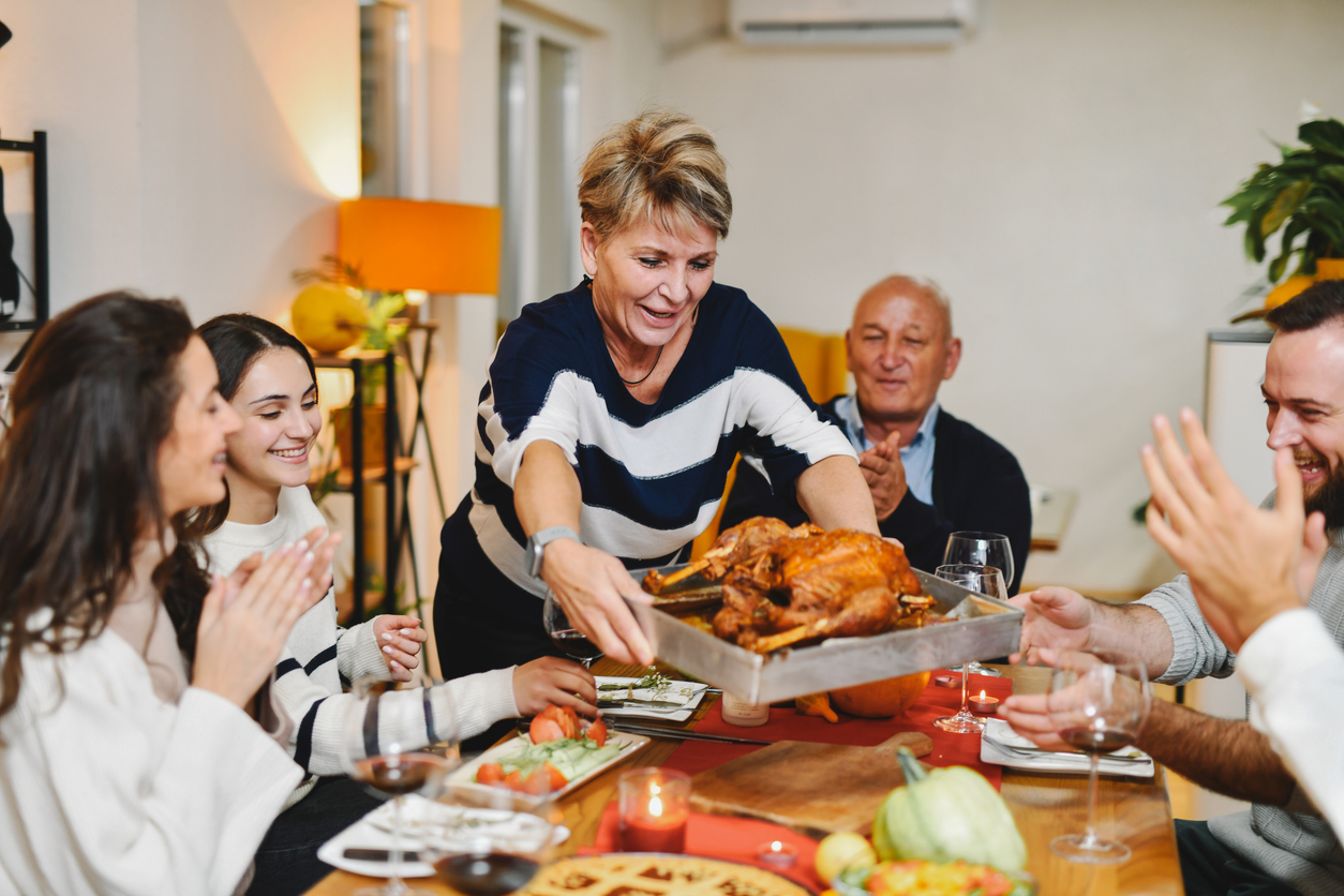 A woman places a roasted turkey on a table as a group of people at a dinner party applaud.