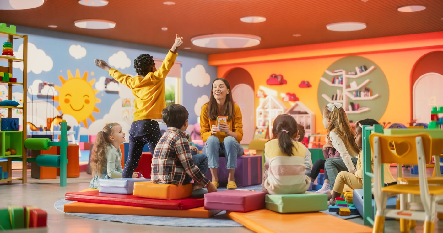 A teacher sits on a floor cushion holding a flashcard for a group of young children in a brightly colored classroom that serves as a budget-friendly alternative to cruise ship charges.