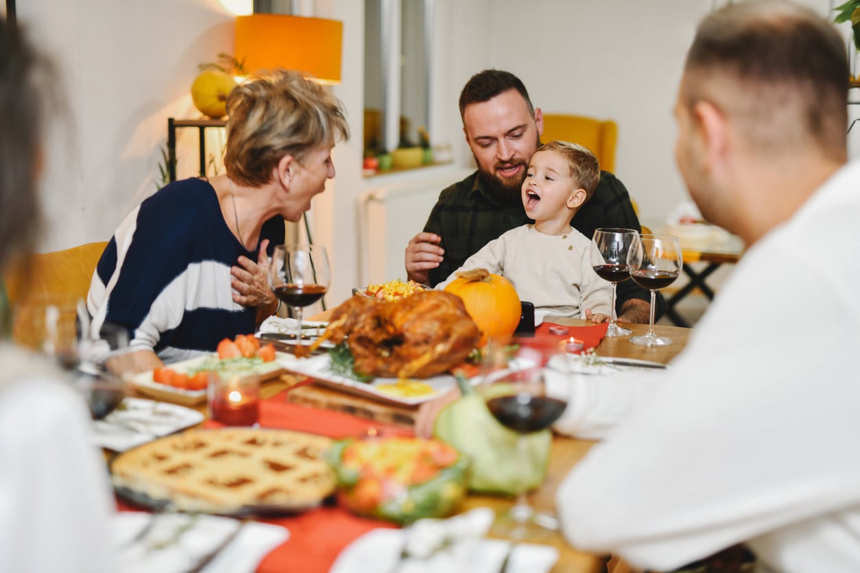 A family sits around a dining table spread with a turkey and other dishes, with a man laughing while holding a young boy who also has his mouth open.