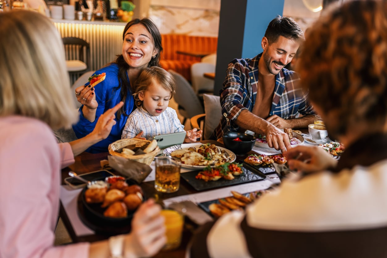 A group of adults and a young child sit at a restaurant table filled with plates of food, discussing menu tips.