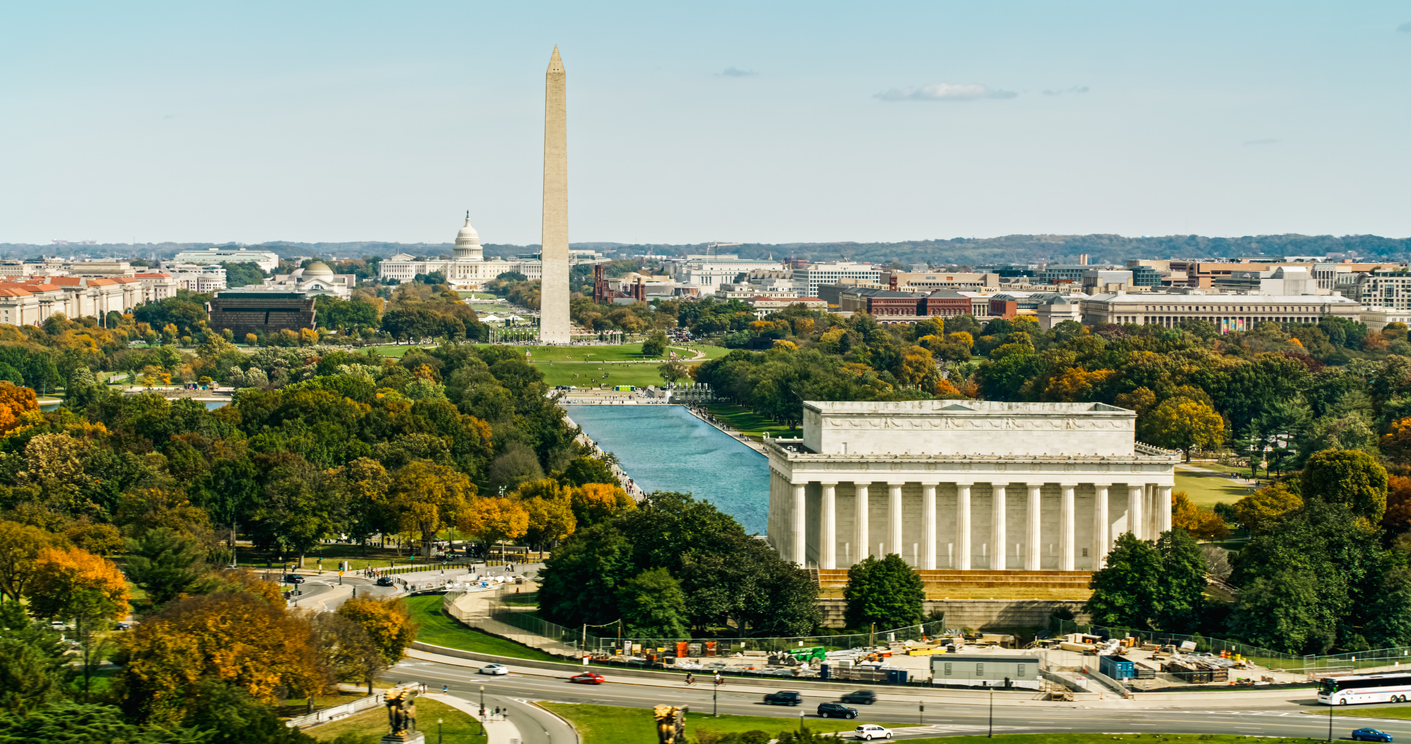 Aerial view of Washington, D.C. showing the Lincoln Memorial, Reflecting Pool, Washington Monument, and U.S. Capitol surrounded by trees and city buildings on a clear day.
