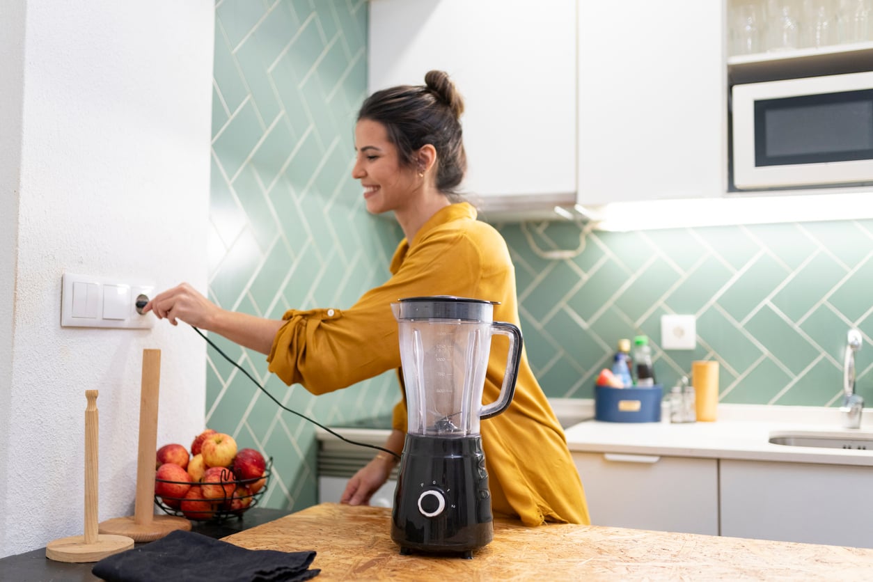 A smiling woman in a yellow shirt plugs a black blender into a wall outlet in a kitchen.
