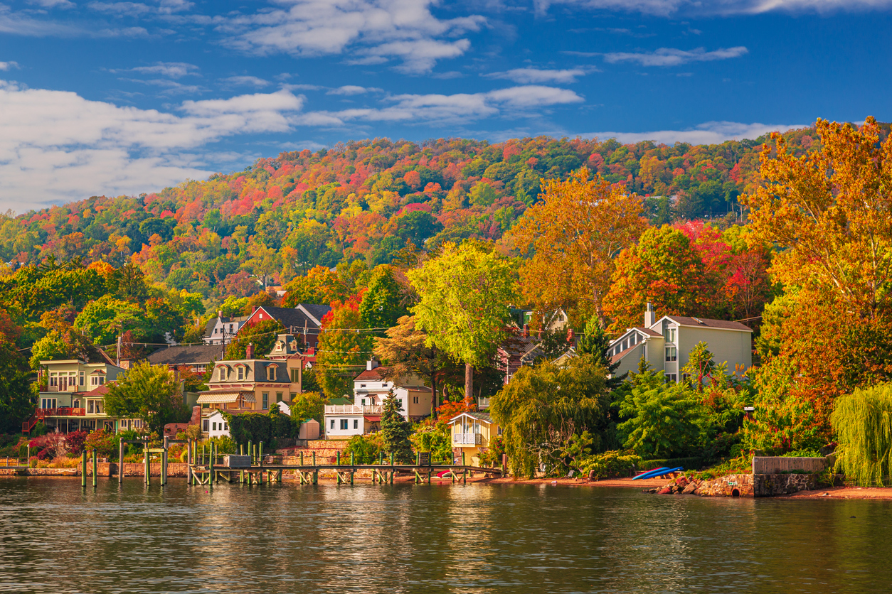 Houses tucked along a lakeshore backed by a hill covered in fall foliage, one of many scenic places to hike in the fall.