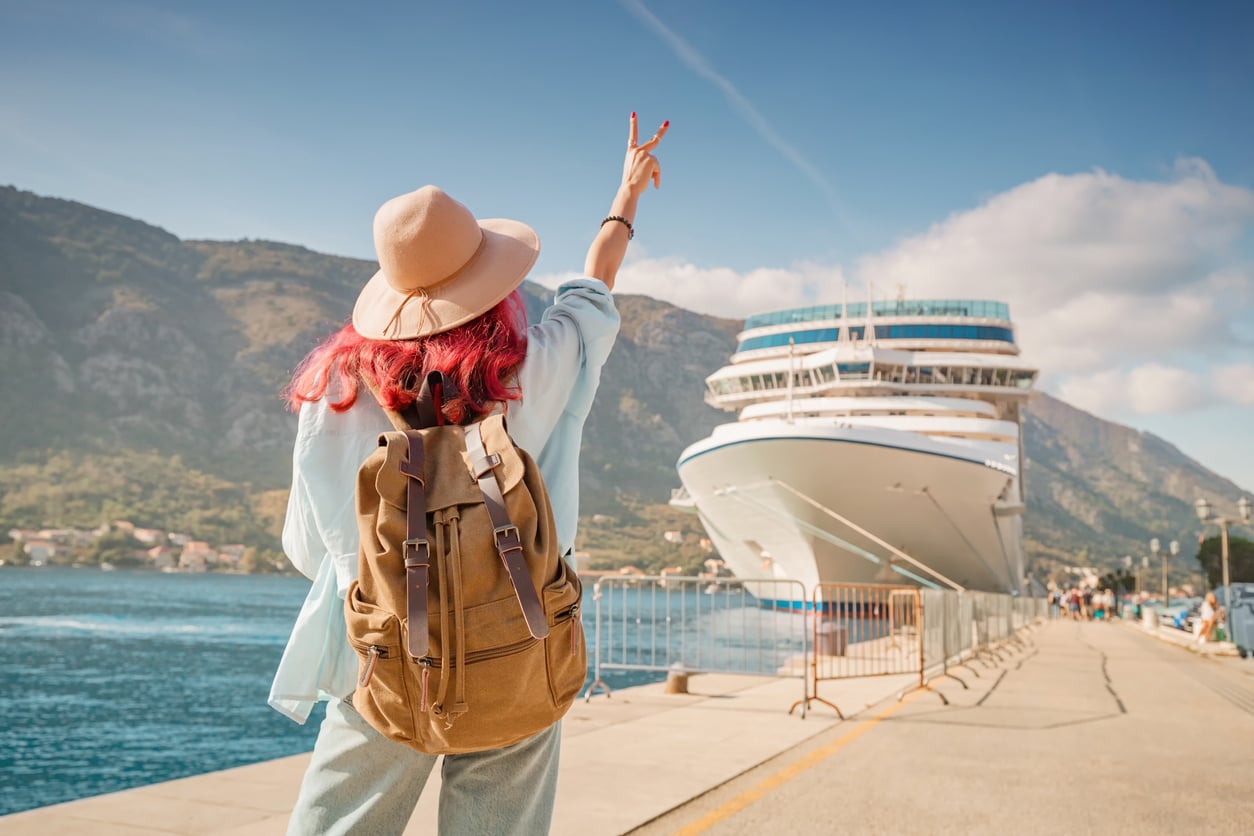 A person with a backpack and hat stands on a pier, making a peace sign toward a large cruise ship docked in a harbor.