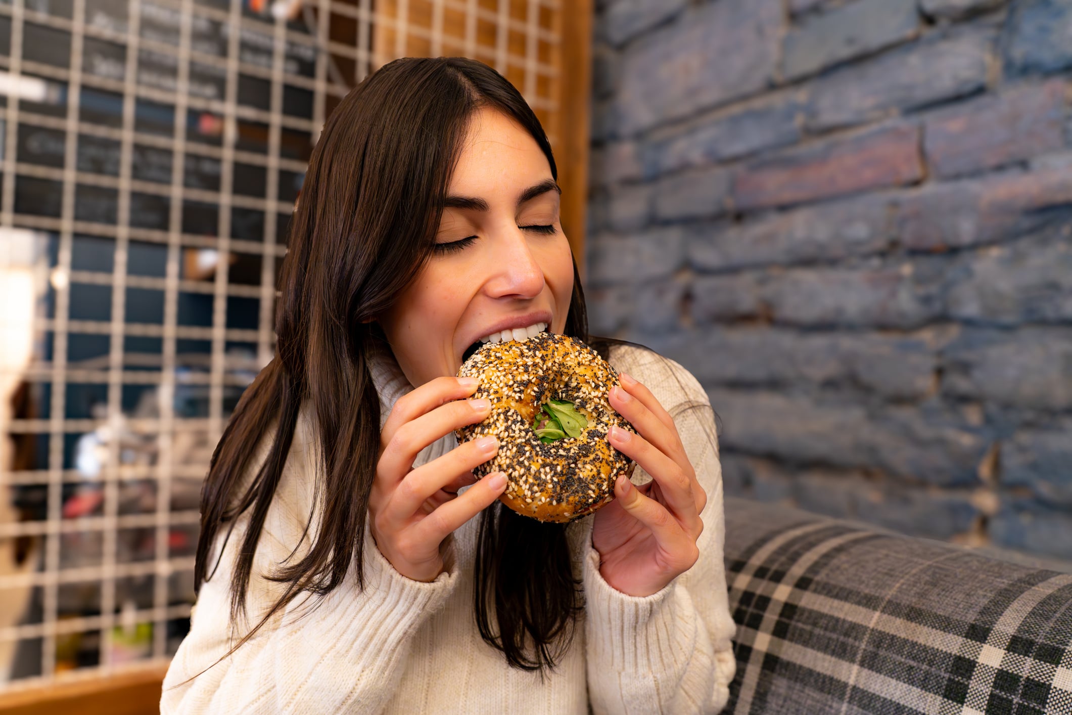 A woman with long dark hair wearing a white sweater takes a bite of an everything bagel for breakfast.