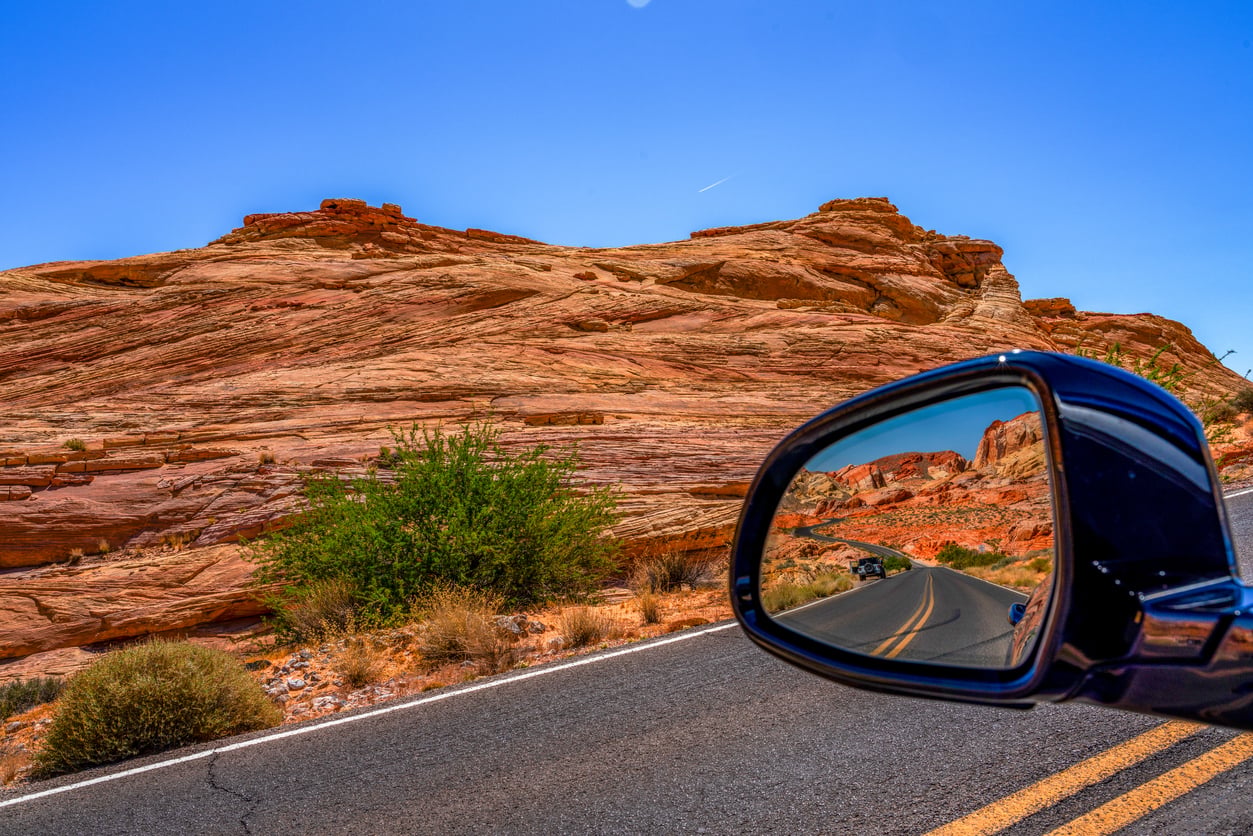 View of a winding road and red rock formations seen through a car’s side mirror.