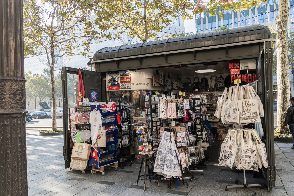 A sidewalk kiosk in Paris displaying souvenir tote bags, postcards, and apparel.