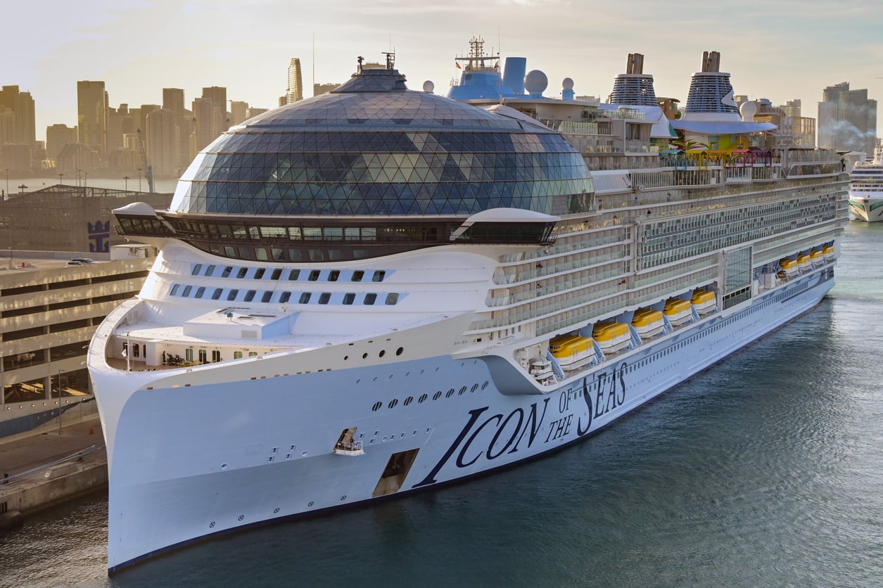 The cruise ship Icon of the Seas, a hub for cruise entertainment, is docked at a port with a city skyline in the background.