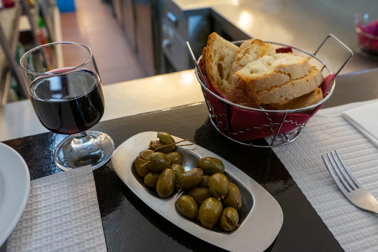 A glass of red wine, a small metal dish of green olives, and a basket of sliced crusty bread on a restaurant table.
