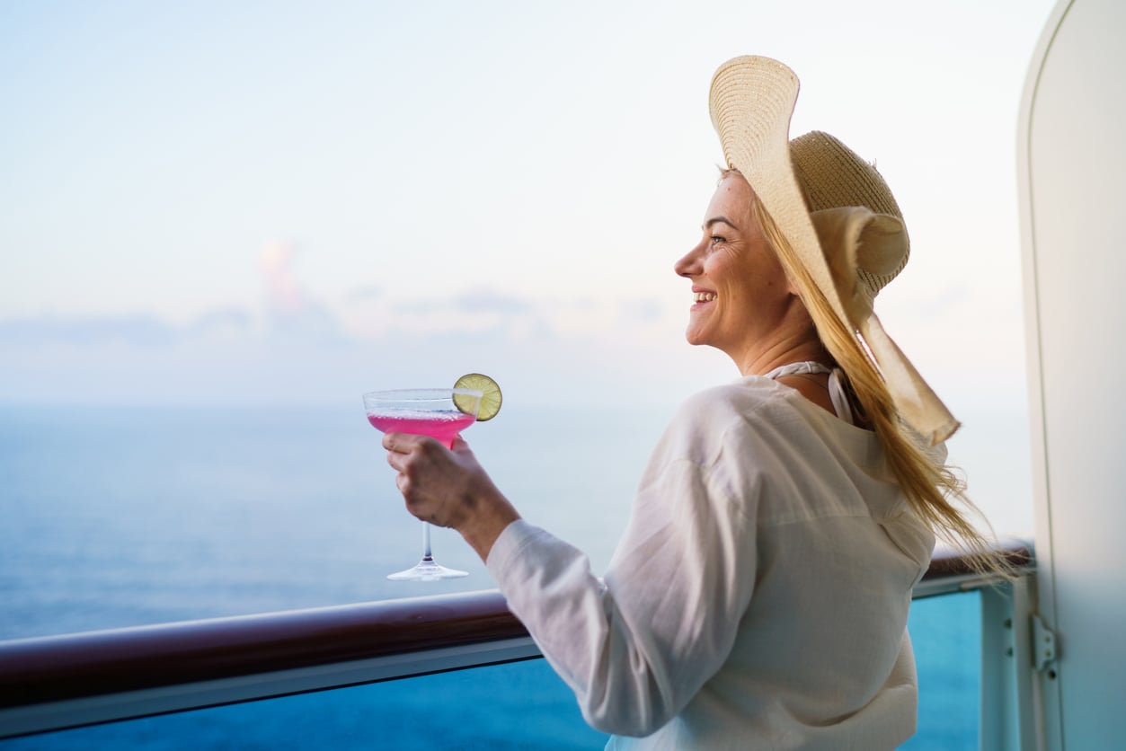 A woman wearing a sun hat stands on a cruise ship balcony, smiling while holding a pink cocktail at sunset.