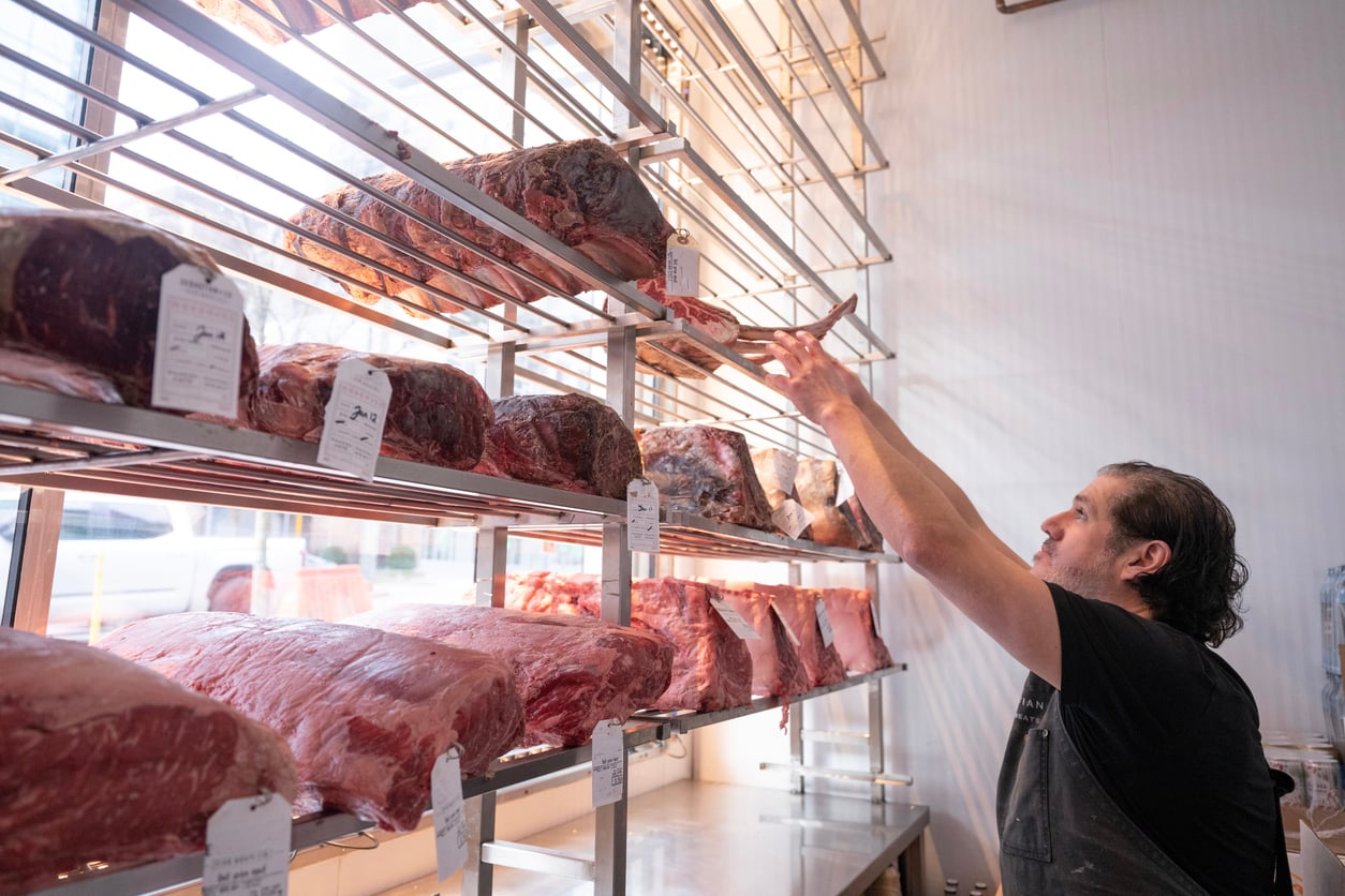 A butcher reaches for a large cut of dry-aged beef on a metal display rack.