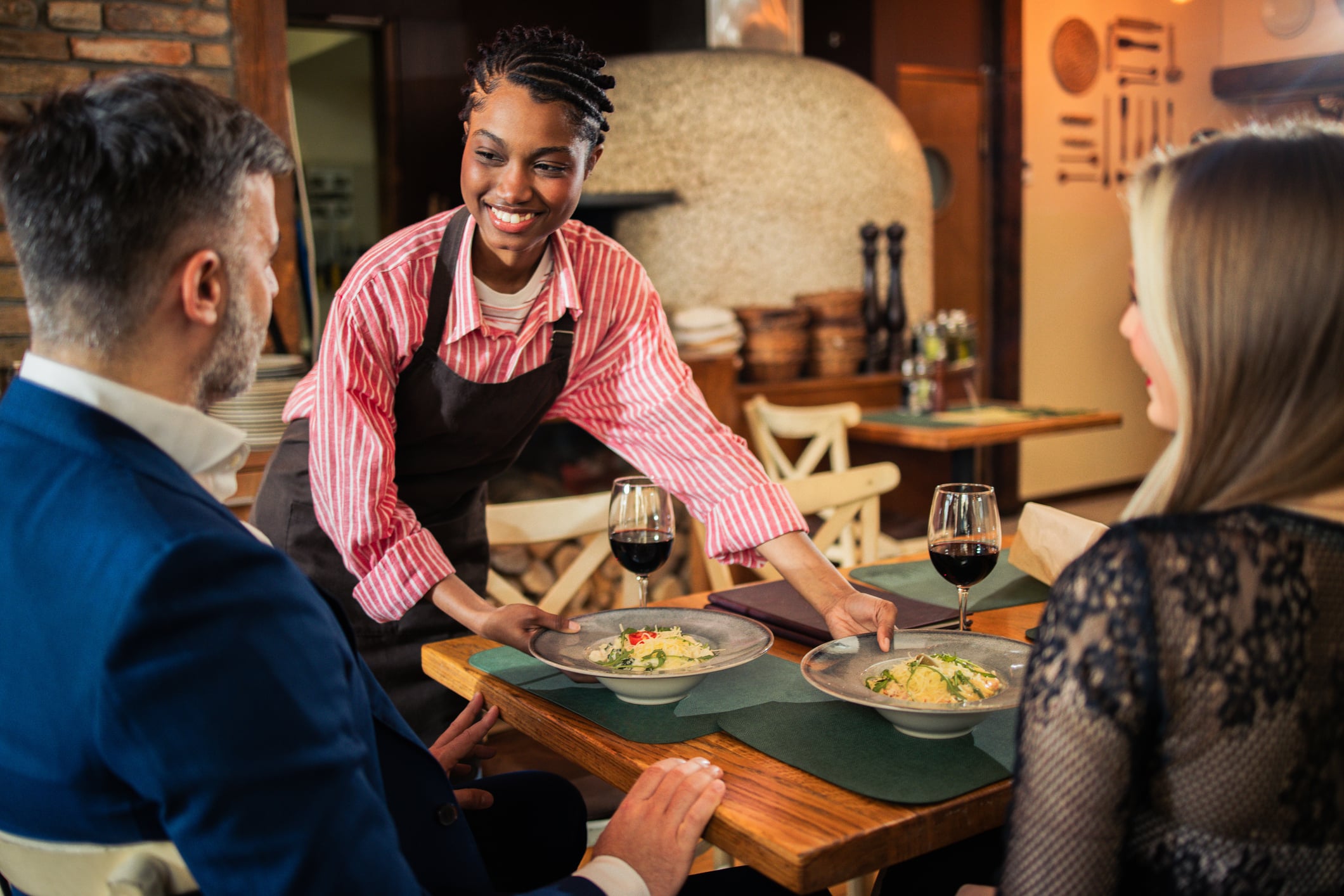 A smiling server in a striped shirt and apron serves plates of pasta to a man and woman at a restaurant table.
