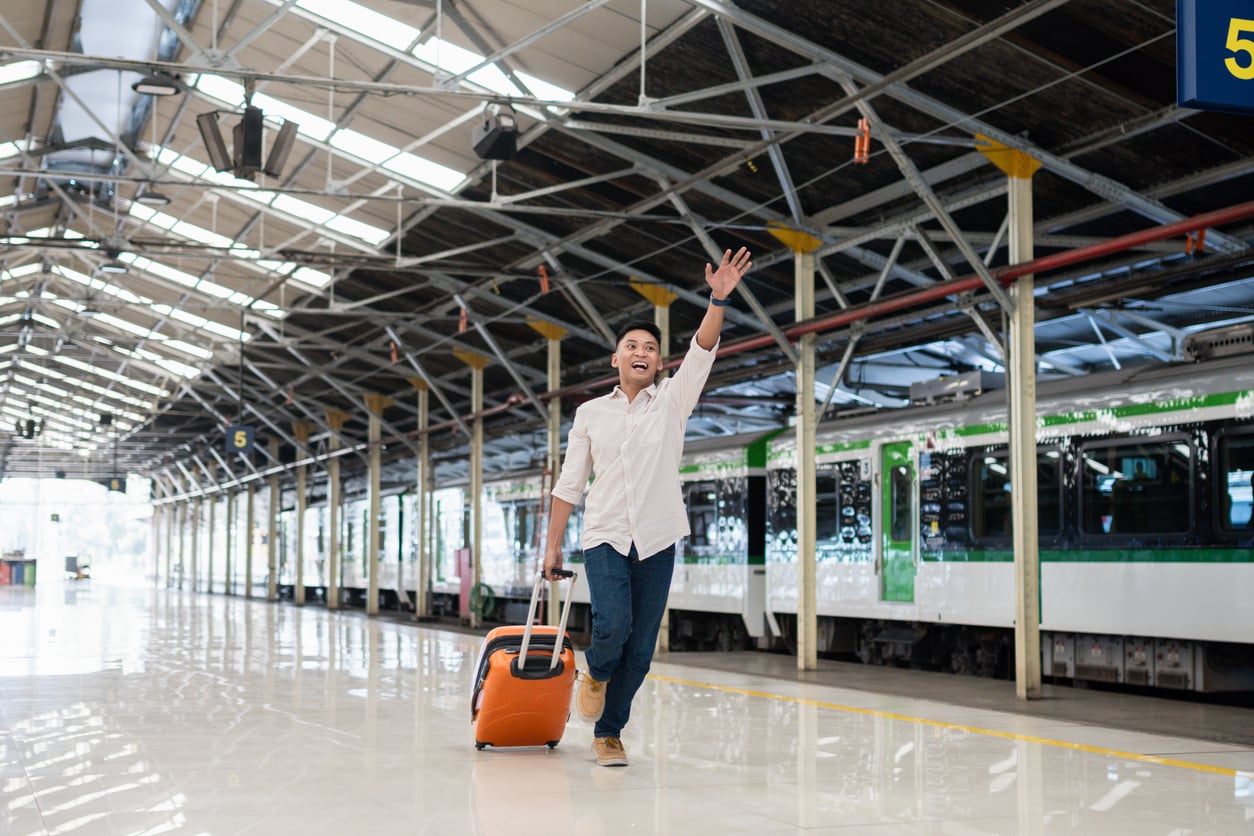 A smiling man in a white shirt and jeans walks along a train platform, pulling an orange rolling suitcase and waving his hand.