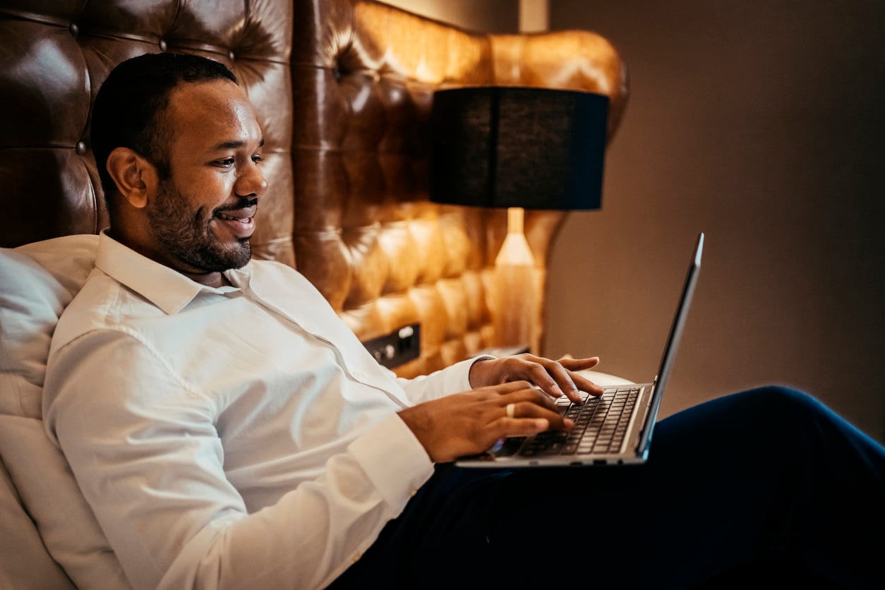 A man in a white dress shirt sits on a bed typing on a laptop while making trip planning decisions.