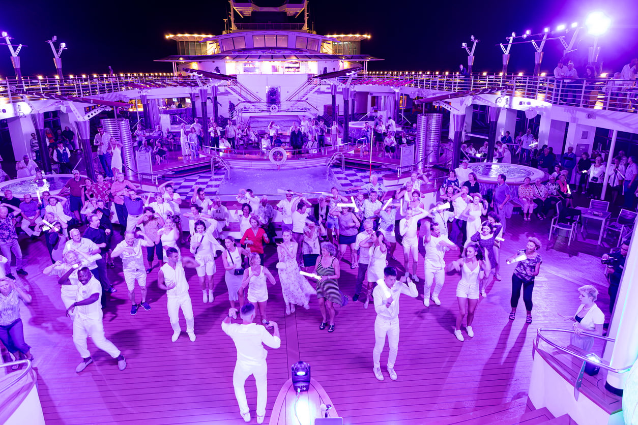 A large group of people in white clothing dancing on a cruise ship deck at night, bathed in purple light.