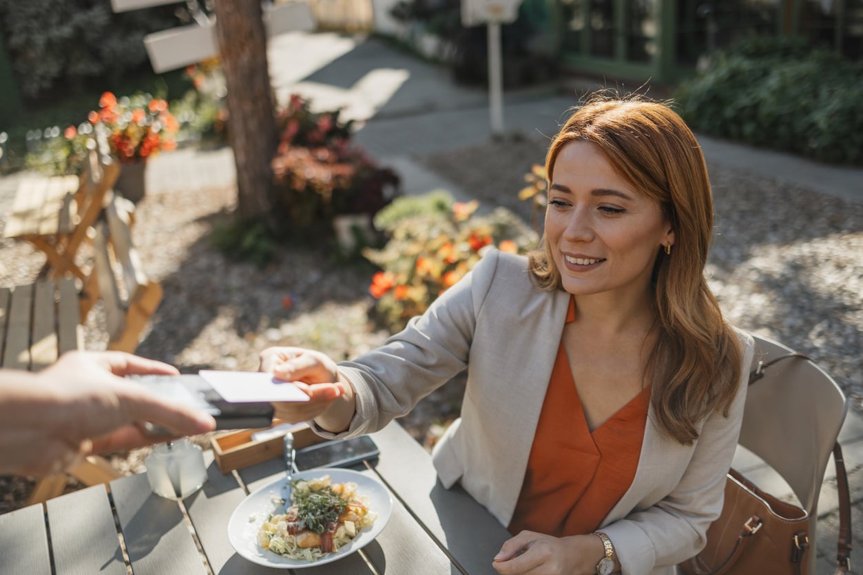 A woman sitting at an outdoor table pays for her meal by tapping a credit card against a portable payment terminal.
