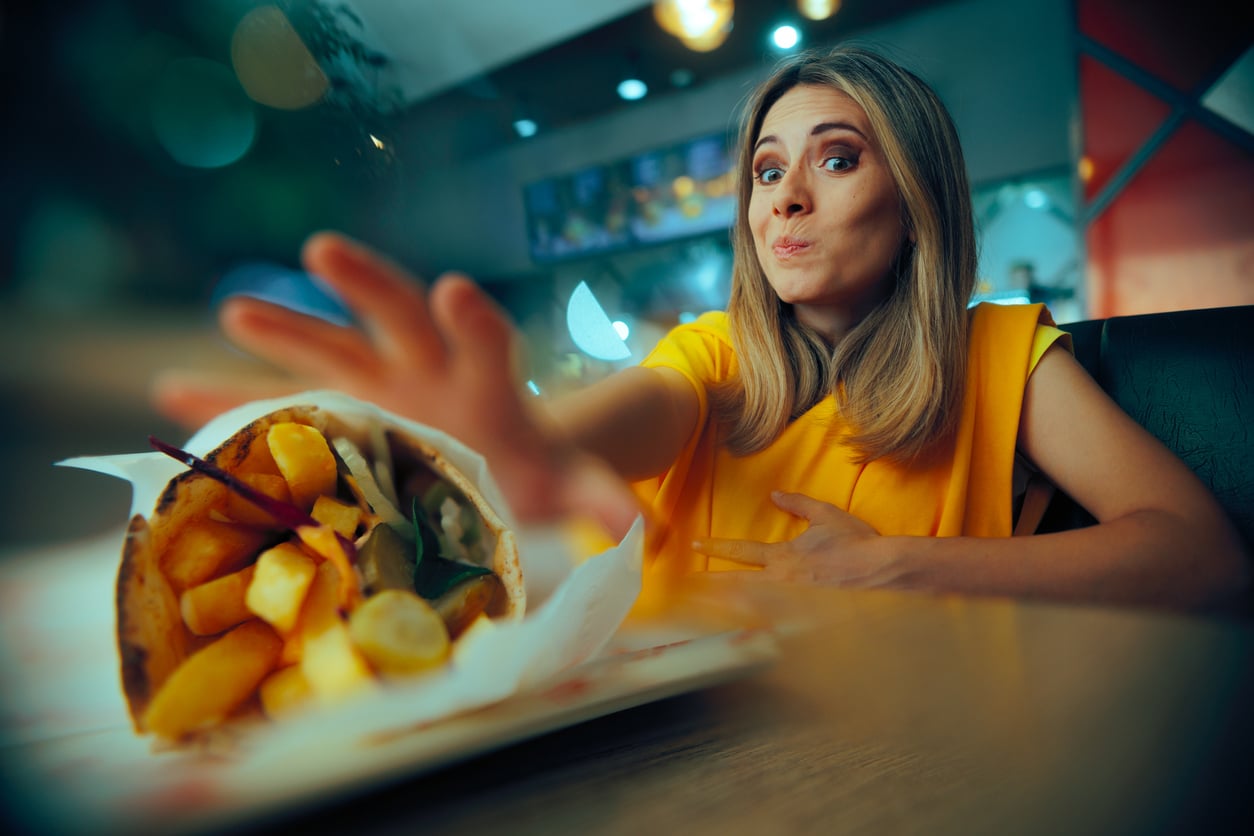 A woman in a yellow top reaches toward a gyro wrap on a restaurant table with a playful, hungry expression.