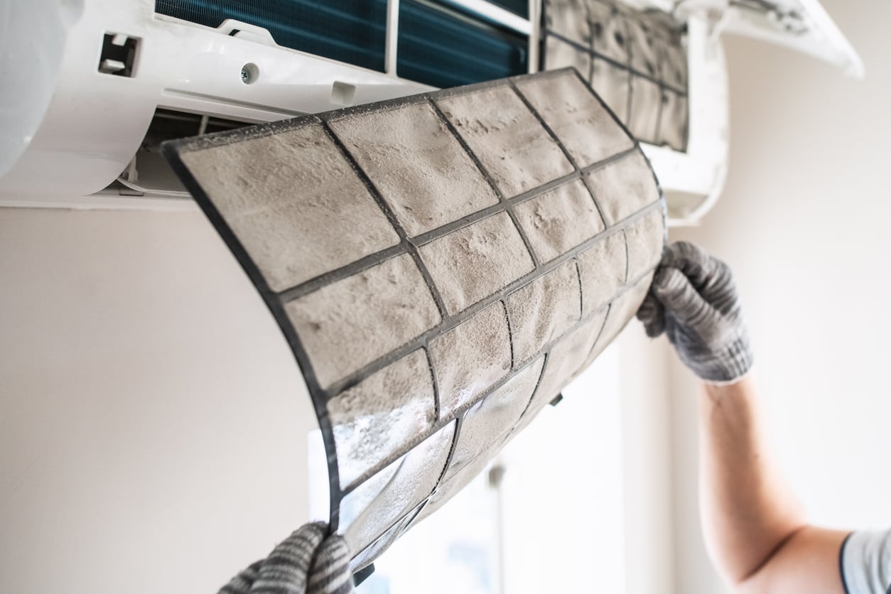 A person wearing gloves removes a dirty, dust-covered air conditioner filter, doing some appliance maintenance from a wall-mounted unit.