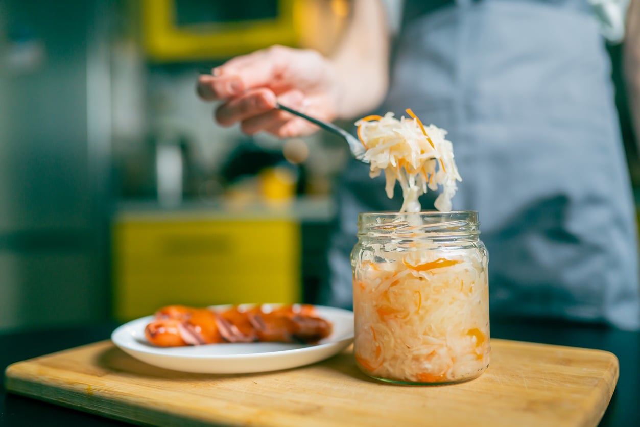 A person uses a fork to lift sauerkraut from a glass jar, with a plate of sliced sausages nearby on a wooden cutting board.