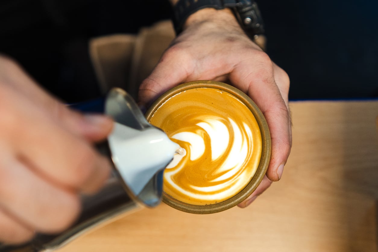 A barista pouring steamed milk into a ceramic cup to create latte art, a relaxing experience often included in cruise ship charges.