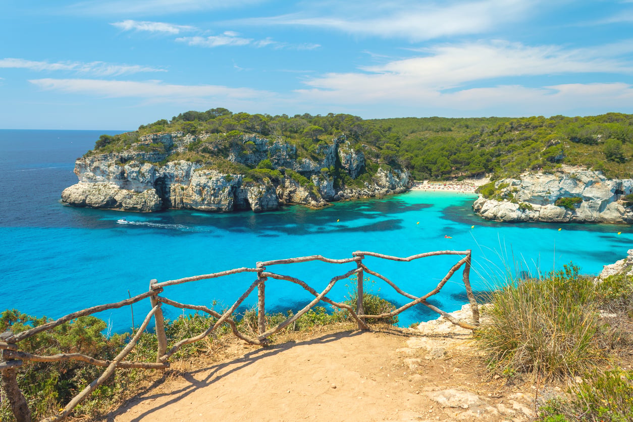 A turquoise cove surrounded by rocky cliffs and pine trees, viewed from a dirt path with a rustic wooden fence.