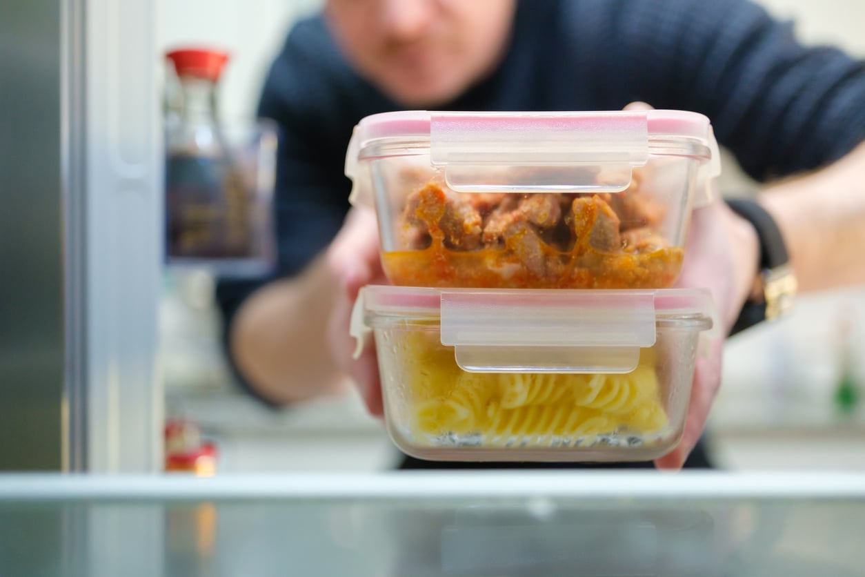 A person holding two stacked glass food containers in front of an open refrigerator.