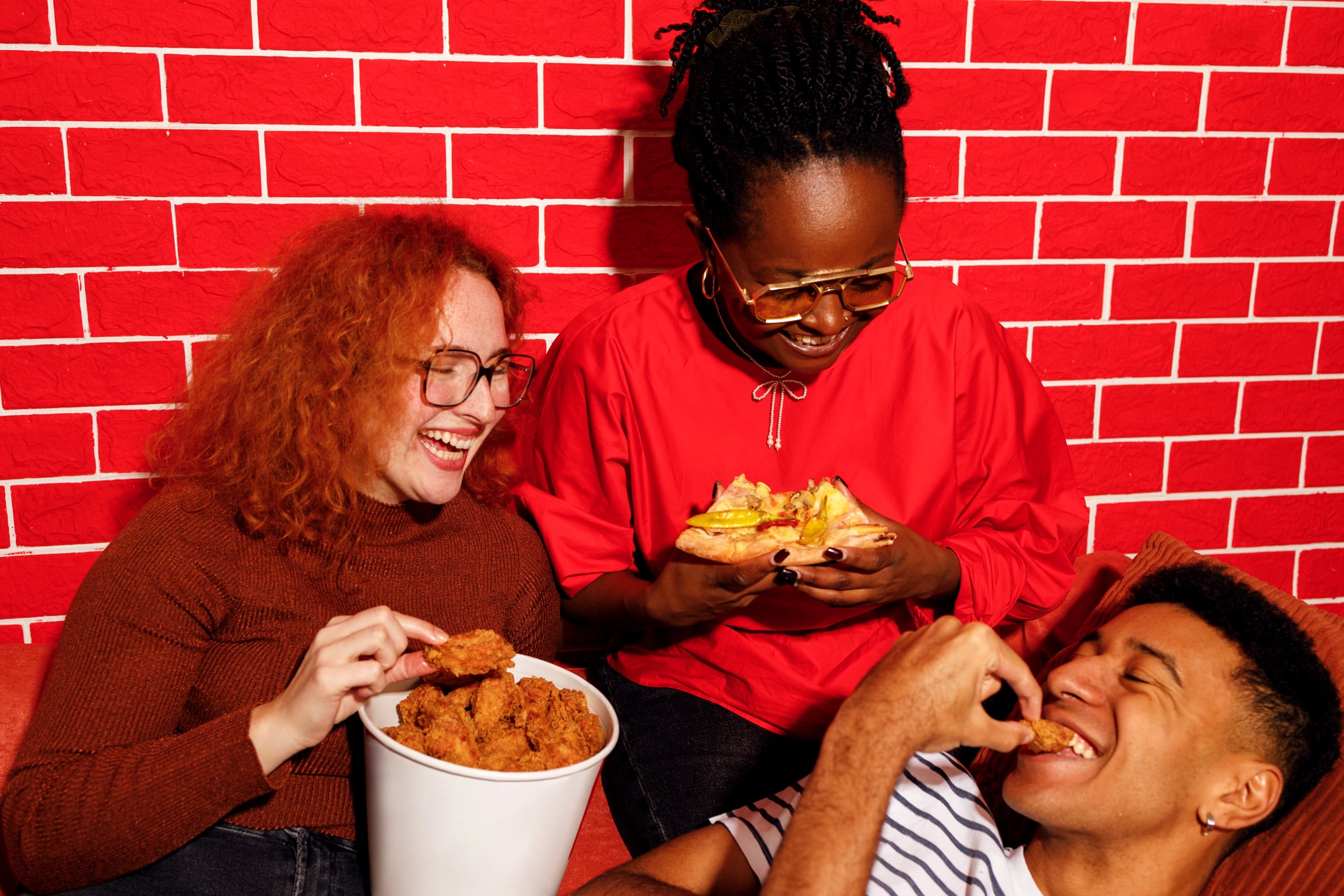 Three friends laughing while eating breakfast foods like fried chicken and pizza in front of a red brick wall.