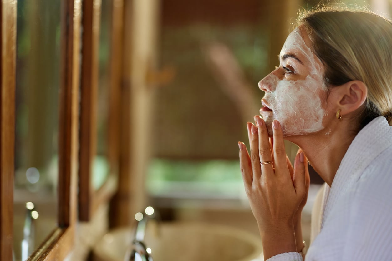 A woman in a white robe looking in a mirror while applying a facial mask.