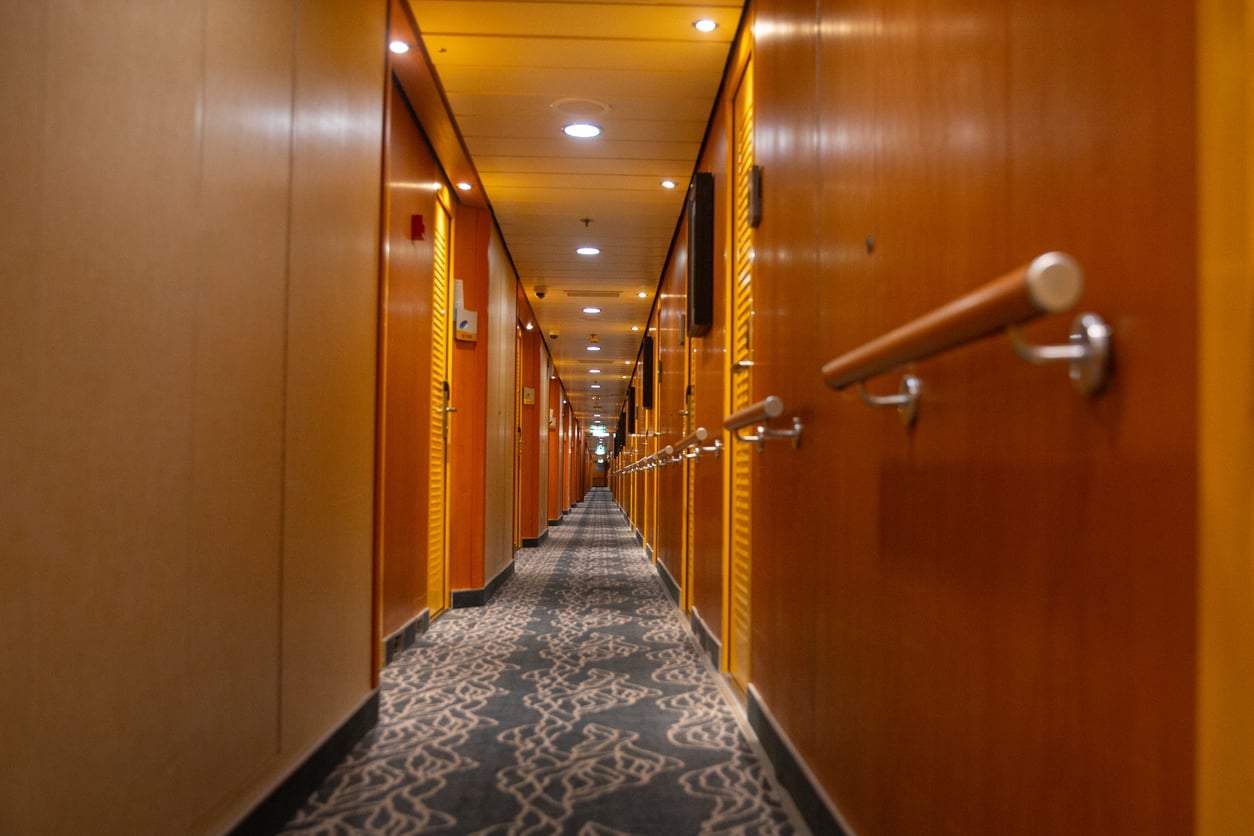 Long cruise ship hallway with patterned carpet, wooden doors, and wall-mounted handrails.