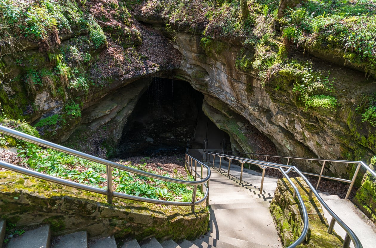 Concrete stairs with metal railings descend into the opening of a moss-covered cave, a popular site among local tourist attractions.
