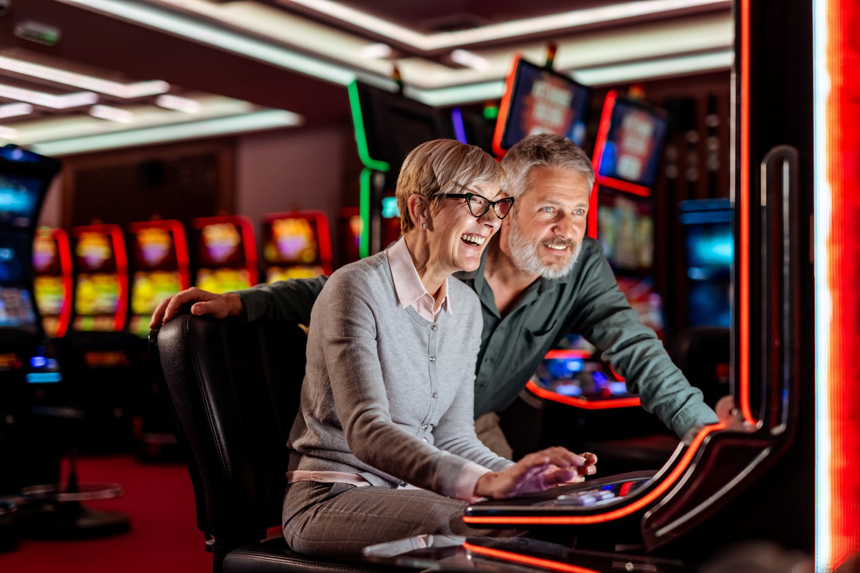A smiling woman and a man looking over her shoulder at a lit-up slot machine in a casino.