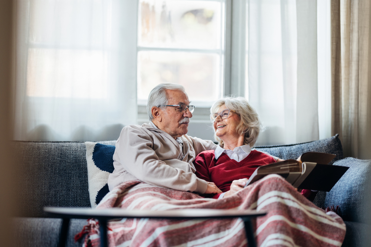 An elderly couple sits together on a sofa under a blanket, smiling and looking at a photo album.