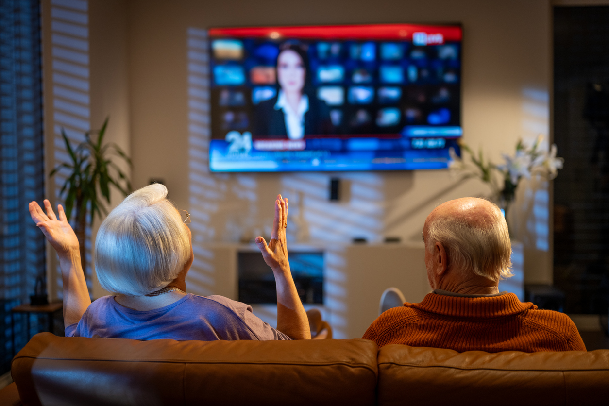 An older couple sits on a sofa watching a news broadcast on a wall-mounted television.