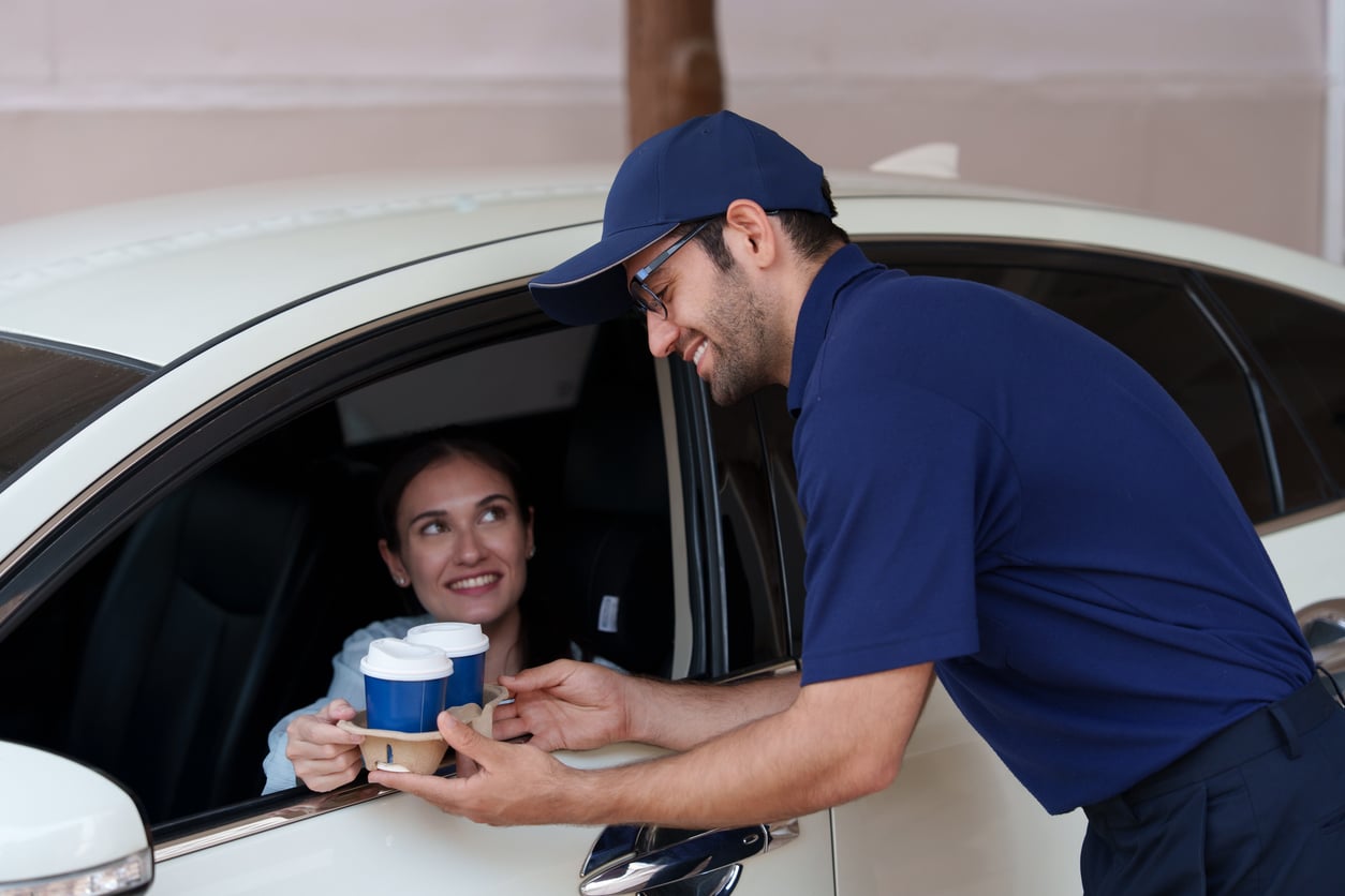 A smiling delivery worker in a blue uniform hands a tray with two coffee cups to a woman sitting in the driver’s seat of a car.