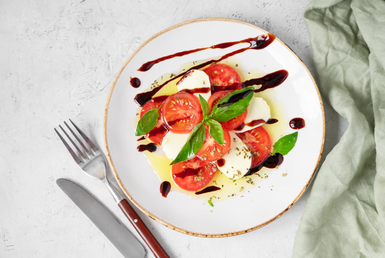 Caprese salad of sliced tomatoes and mozzarella garnished with fresh basil, olive oil, and balsamic glaze on a white plate with a fork and knife.