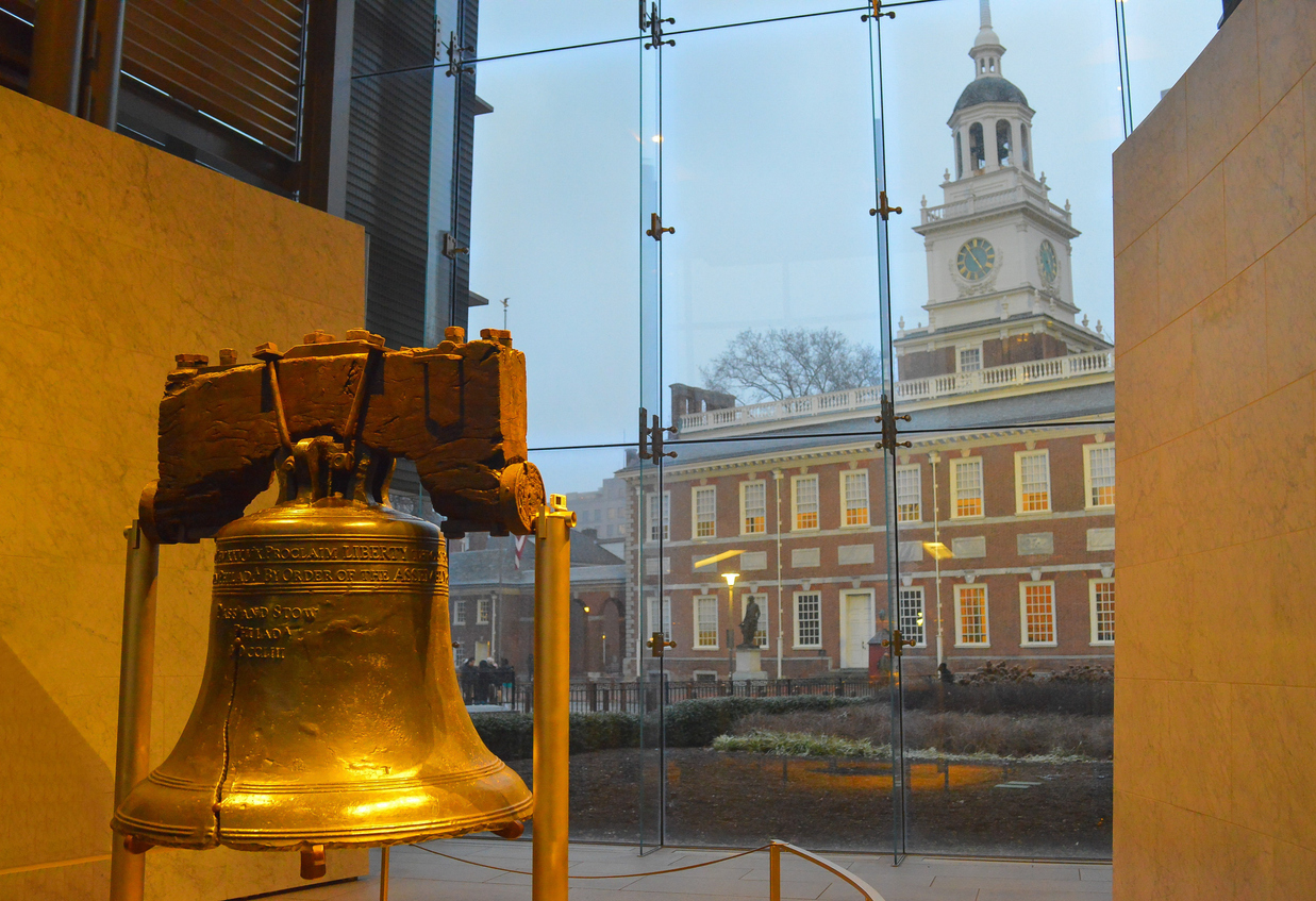 The Liberty Bell on display inside a building, with Independence Hall visible through a large glass window behind it.