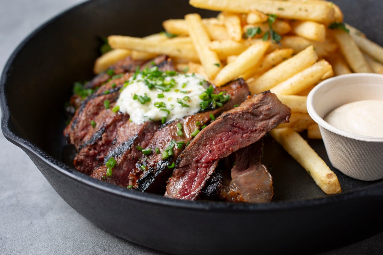 Sliced steak topped with herb butter and chives, served with french fries and a side of dipping sauce in a cast iron skillet, a perfect addition to any restaurant menu.