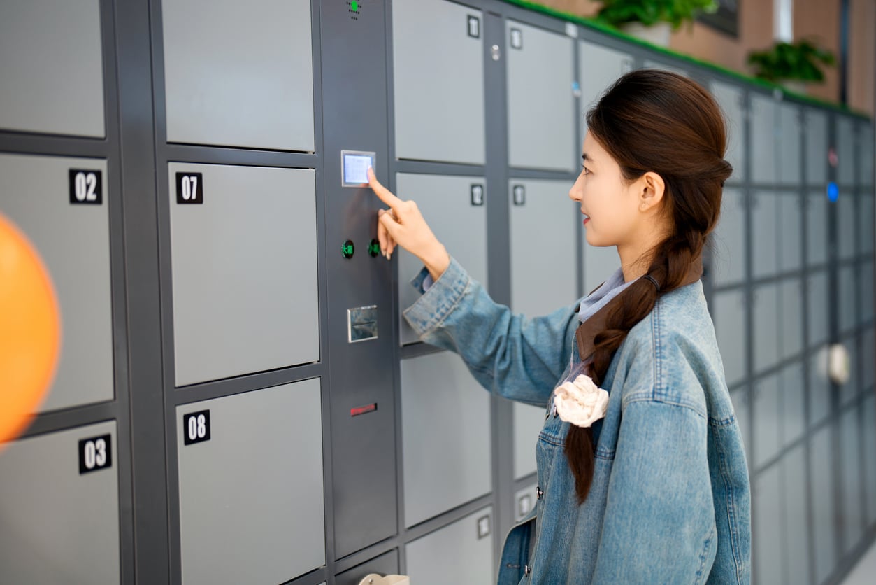 A woman in a denim jacket touches the digital screen of a numbered metal storage locker.