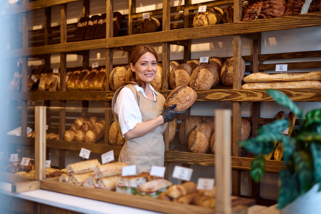 A smiling bakery employee in an apron holds a loaf of sourdough bread in front of shelves filled with various artisanal breads.