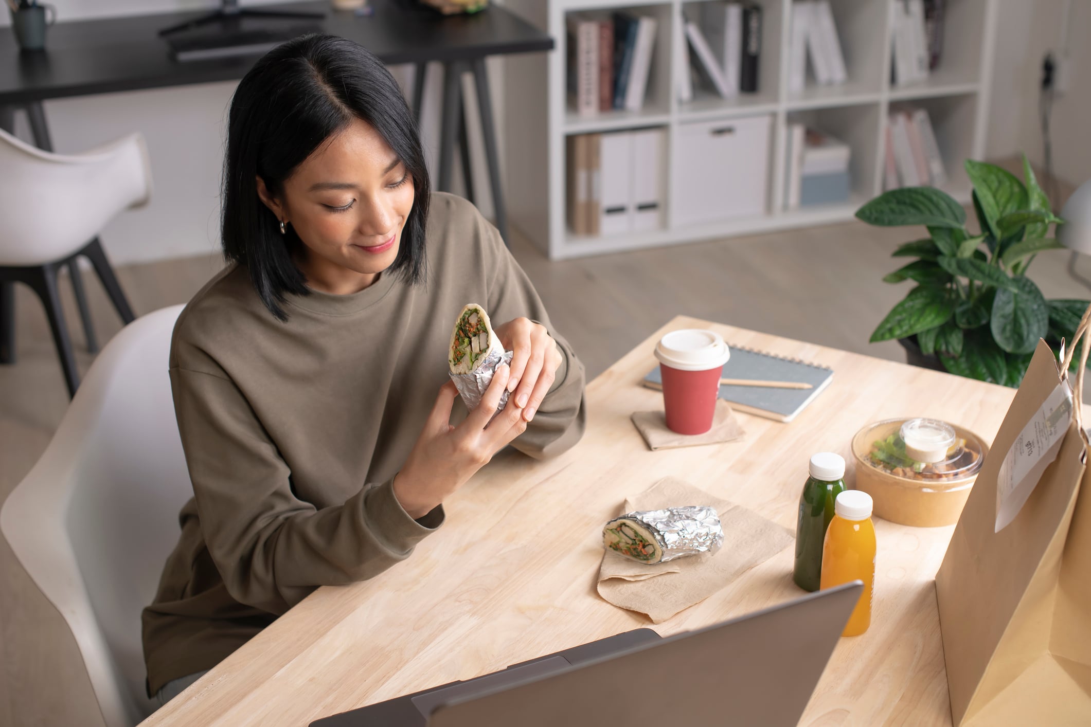 A woman sits at a desk with a laptop, looking at a foil-wrapped breakfast food held in her hands.