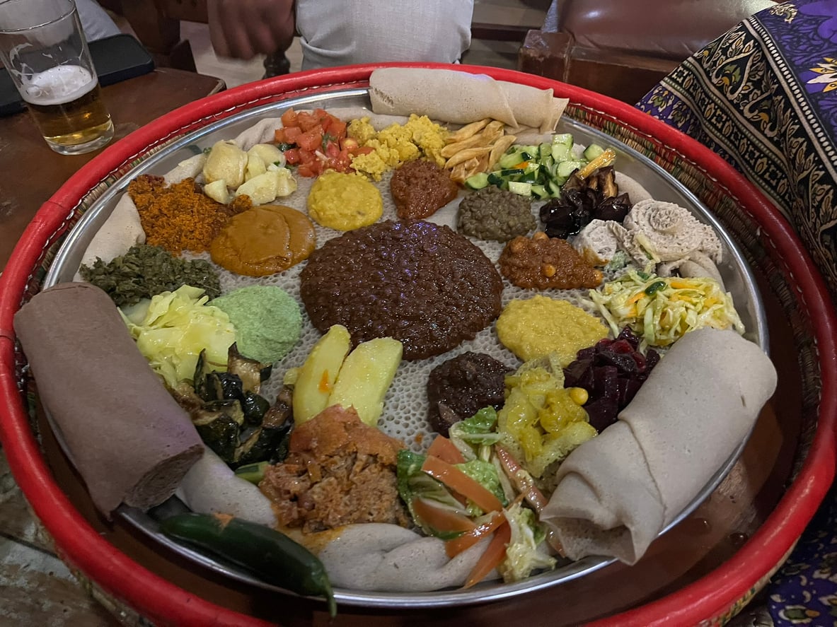 A large circular platter featuring an assortment of Ethiopian stews, vegetables, and salads served atop a layer of injera flatbread, with additional rolled injera on the side.