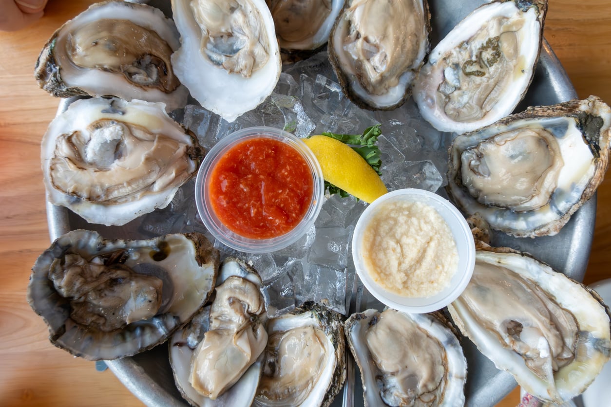 A metal platter of raw oysters on ice, served with small cups of cocktail sauce and horseradish, and a lemon wedge.
