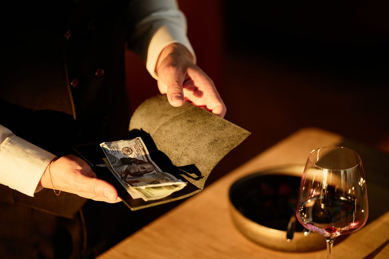 A server in a vest holds a check presenter containing a hundred-dollar bill at a table set with a wine glass.