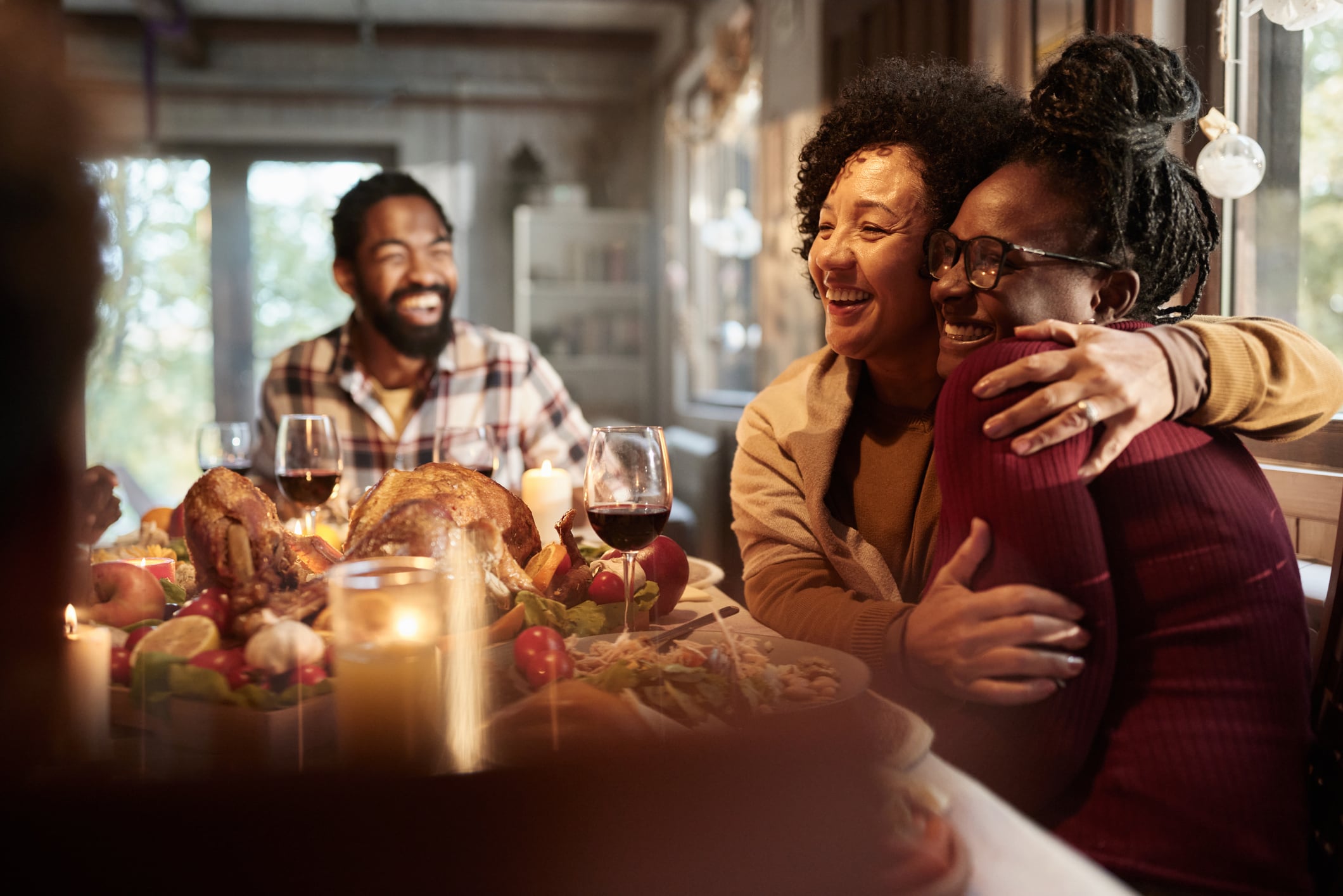 Two women embrace warmly at a dinner table while a man laughs in the background.