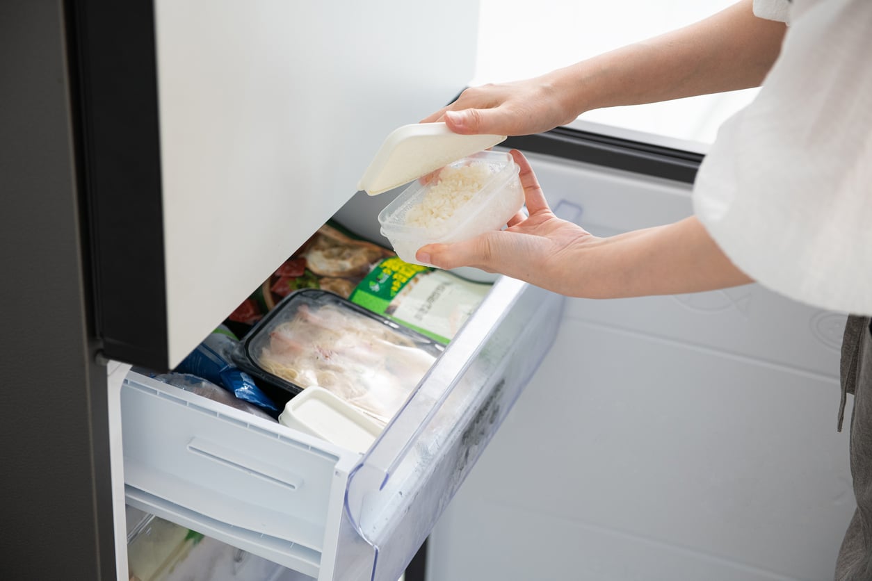 A person holding a small plastic container of cooked rice while taking it out of a freezer drawer.