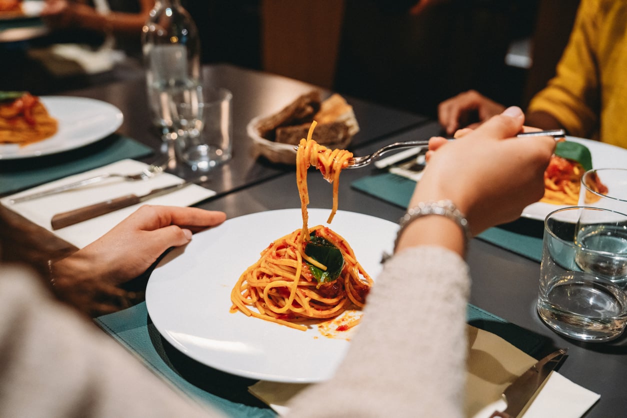 A person uses a fork to lift spaghetti with tomato sauce and a basil leaf from a plate.