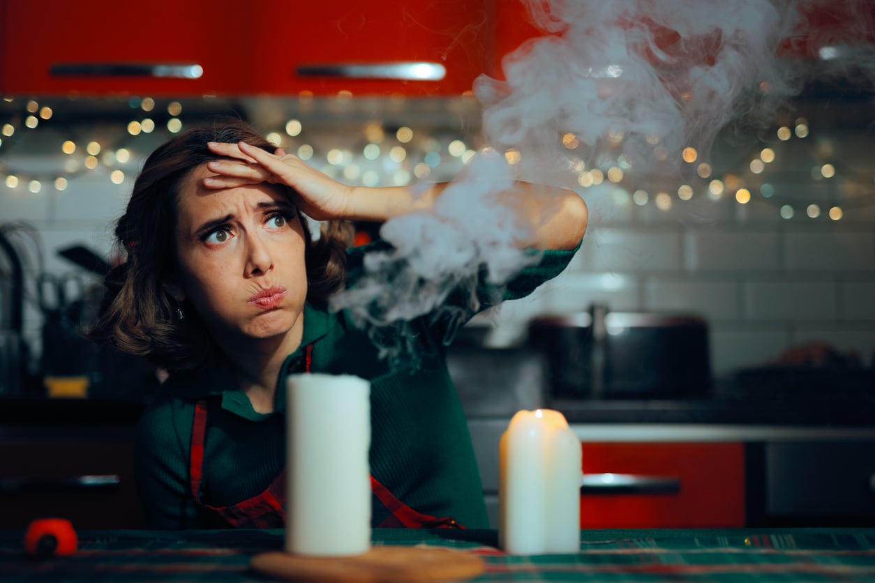A woman in a kitchen looks stressed and holds her forehead while a column of smoke rises from a candle in front of her.