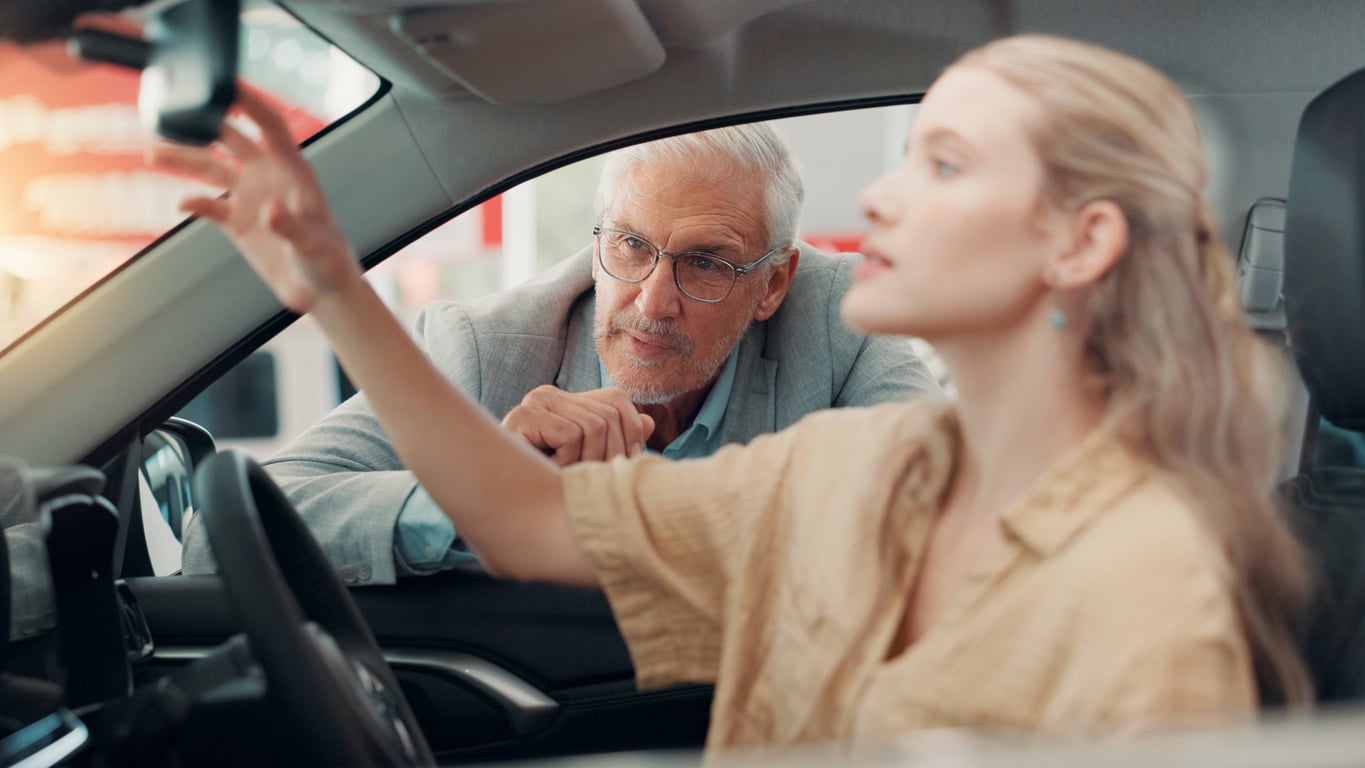 A woman sits in the driver’s seat of a car adjusting the rearview mirror while a man looks on through the open window.